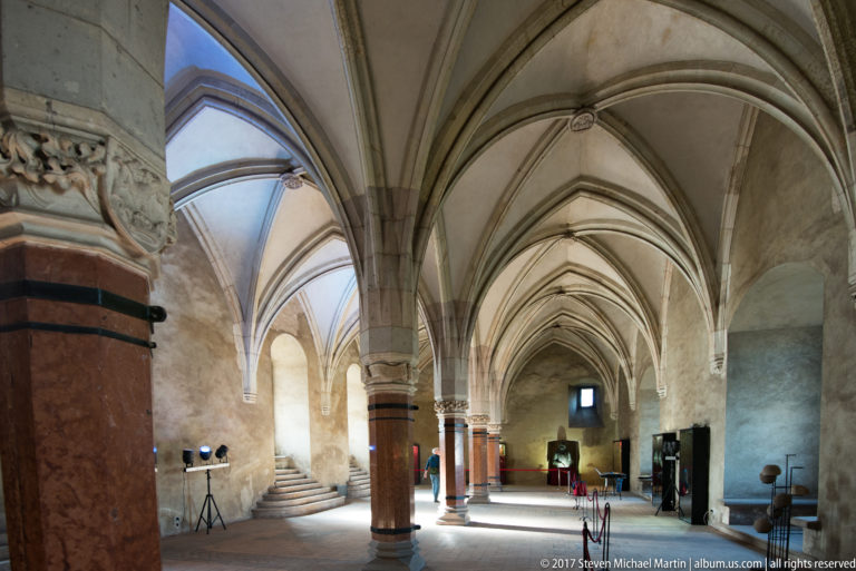 Long view through Corvin Castle hall with pointed rib vaults, stone supports, side opening, and museum equipment.