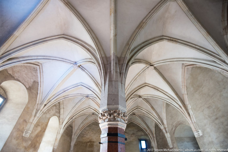 Upward view of intersecting Gothic ribs, pointed vaults, and carved capital inside Corvin Castle.
