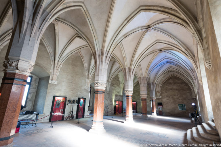 Wide interior view with Gothic rib vaults, stone columns, bright side light, and museum display panels.