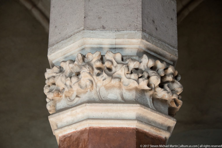 Close-up of a carved stone capital with projecting foliate and mask-like forms above a reddish column.