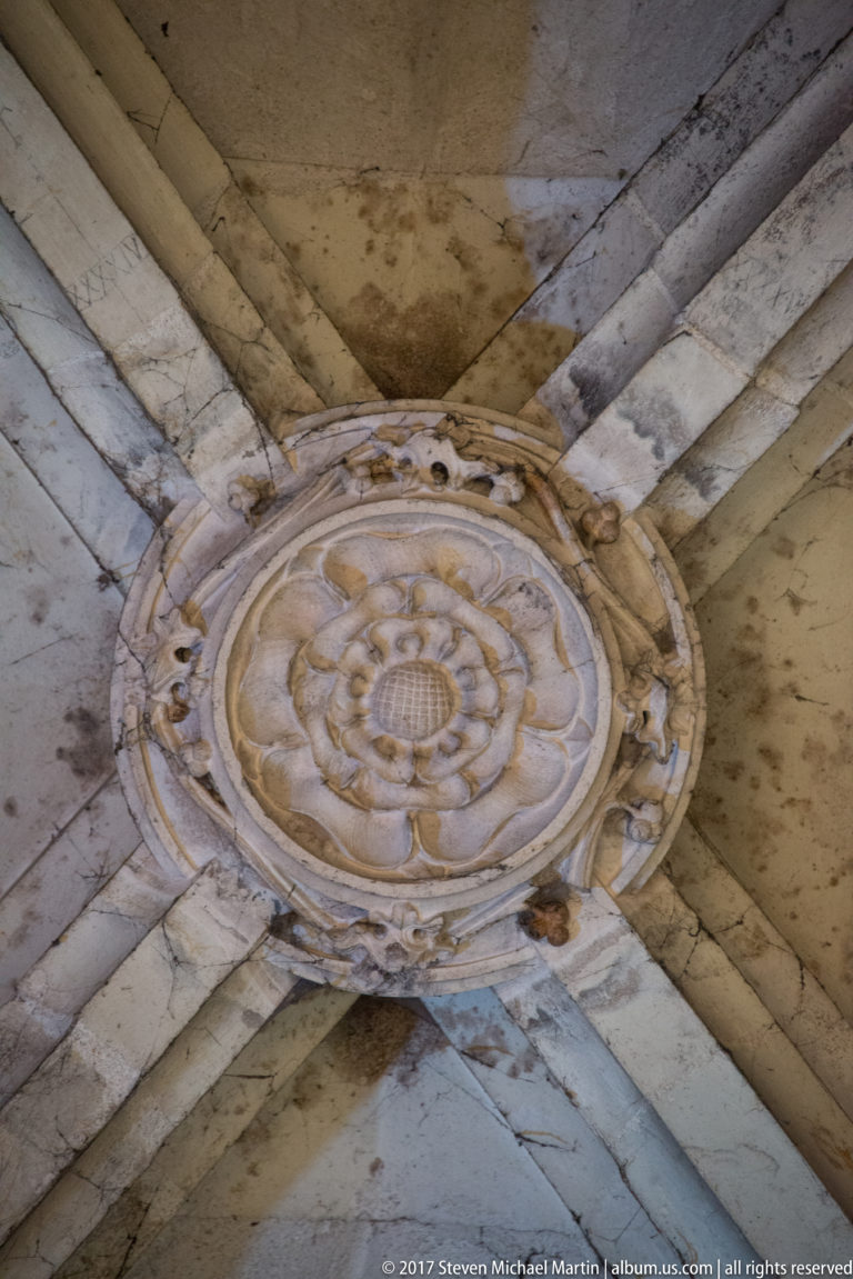 Round carved rosette boss at the intersection of pale stone ceiling ribs inside Corvin Castle.