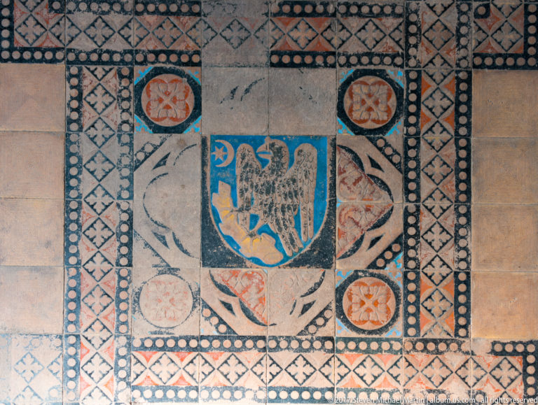 Decorative floor panel with central shield bearing a bird, surrounded by geometric and circular motifs.