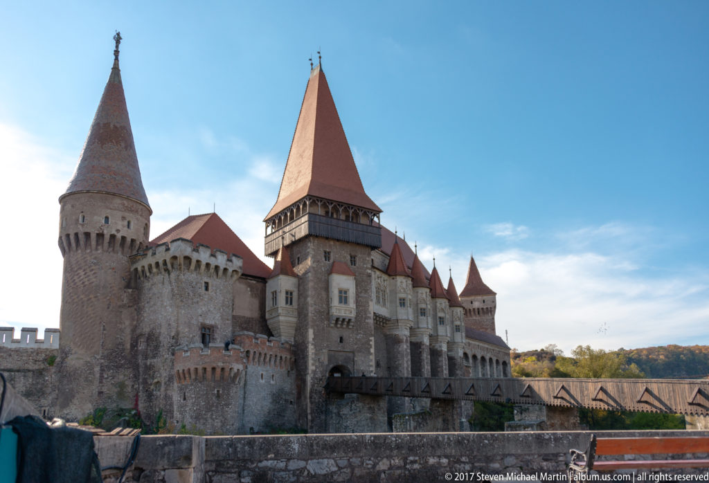 Corvin Castle exterior with round tower, tall roofed gate tower, stone walls, and bridge approach under blue sky.