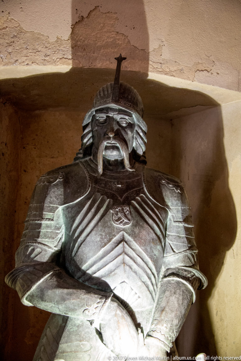 Recumbent armored figure with helmet, plate armor, and folded hands displayed in a castle niche.