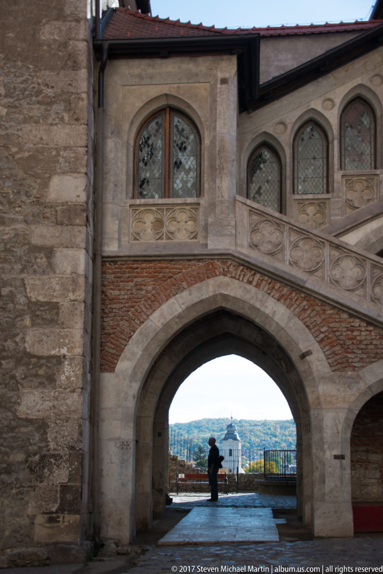 Large courtyard arch framing a visitor and distant landscape beneath Corvin Castle’s upper gallery.