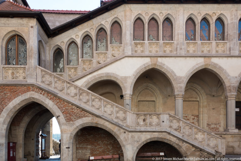 Diagonal stone staircase crossing arches below a decorated upper gallery in Corvin Castle’s courtyard.