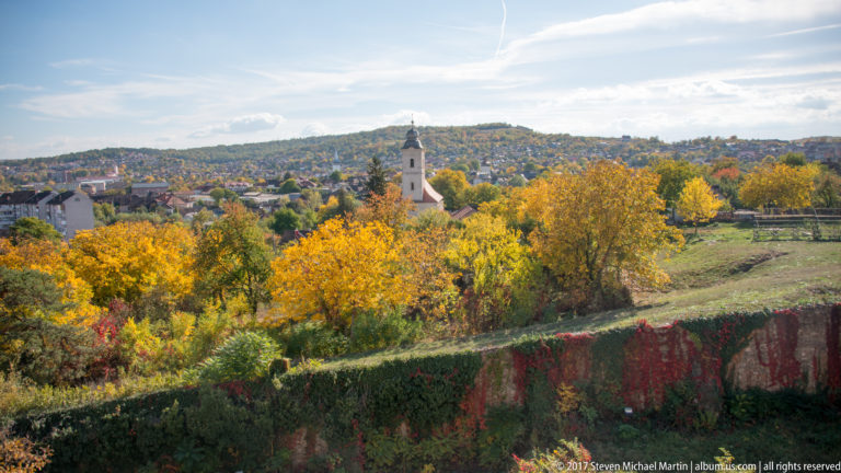Autumn landscape from Corvin Castle with vine-covered wall, trees, town, and a church tower in the distance.