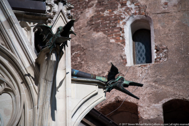Dark dragon-like metal waterspouts projecting from Gothic stonework beside a weathered brick wall and small window.