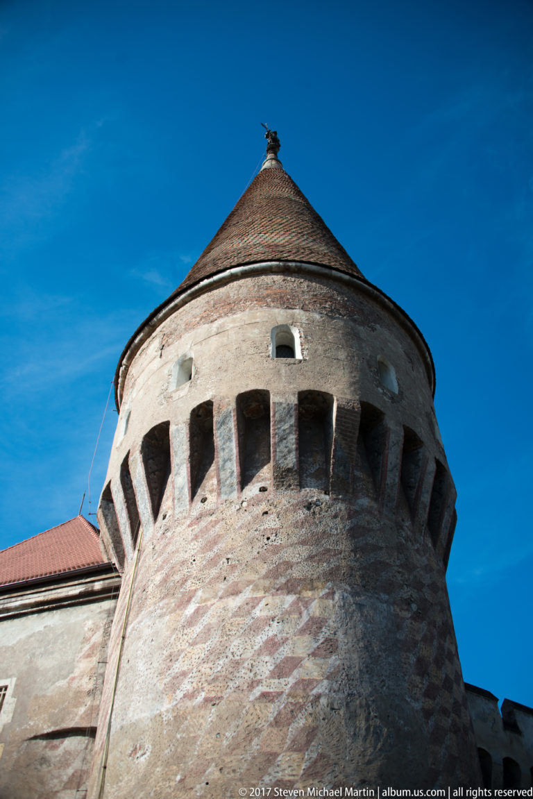 Upper round tower with projecting defensive openings, small windows, and a conical tiled roof against blue sky.