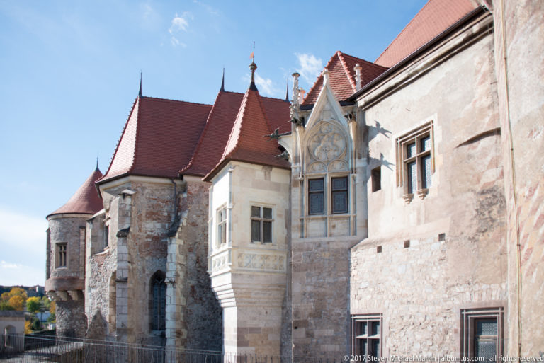 Sunlit Corvin Castle wing with red roofs, projecting bays, and an ornate Gothic window in pale stonework.