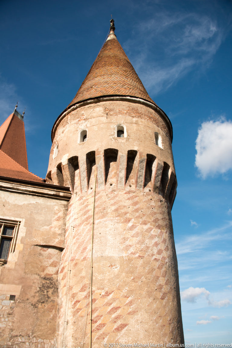 Round Corvin Castle tower with thick masonry, projecting upper defenses, and conical tiled roof under blue sky.
