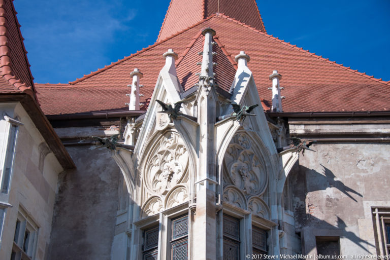 Ornate pointed Gothic window with pale stone tracery, pinnacles, and sculptural details beneath a red roof.
