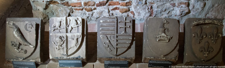 Row of carved stone heraldic fragments with shields and relief emblems displayed against a masonry wall.