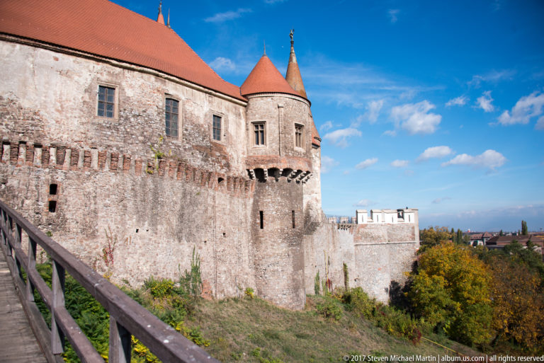 Castle wall and round roofed tower beside the wooden approach bridge under a blue autumn sky.