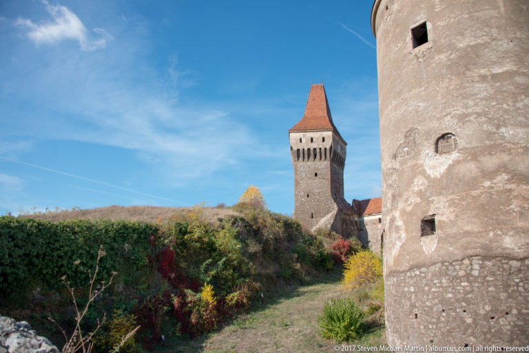 Square defensive tower beyond vegetation with part of a large round tower in the foreground.