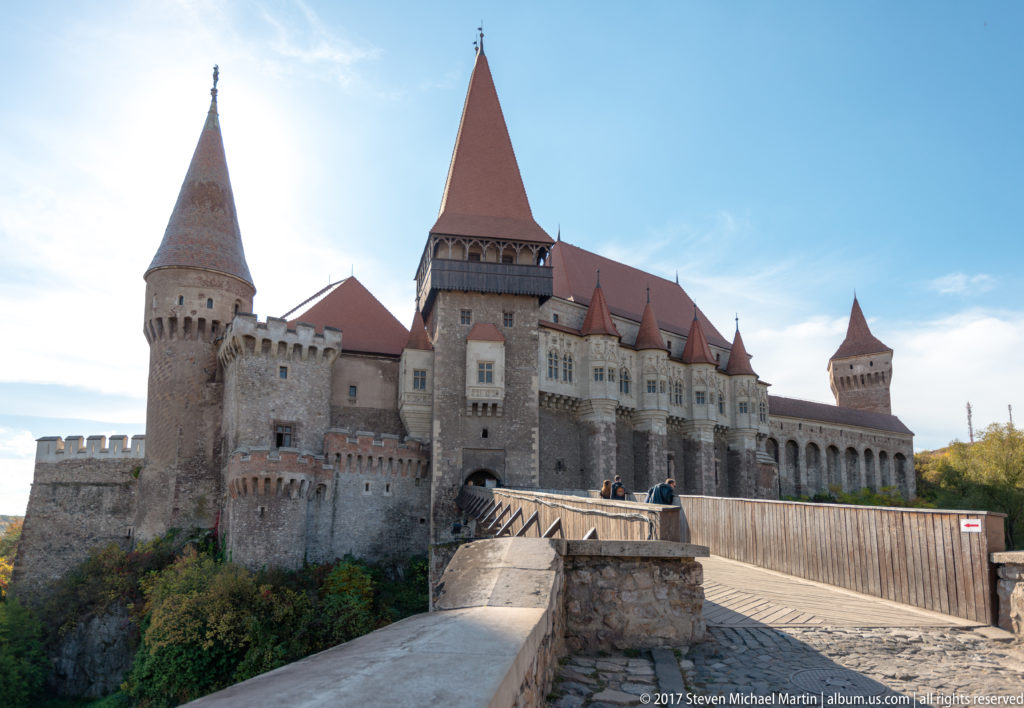 Bridge leading toward Corvin Castle’s central gate tower, flanked by stone towers and red-roofed wings.