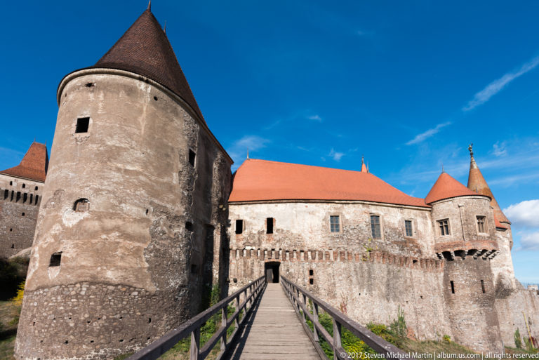 Wooden bridge leading toward Corvin Castle between a round tower and long red-roofed wing.