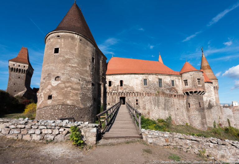 Wide view of Corvin Castle with bridge, round tower, long wing, low stone walls, and distant square tower.