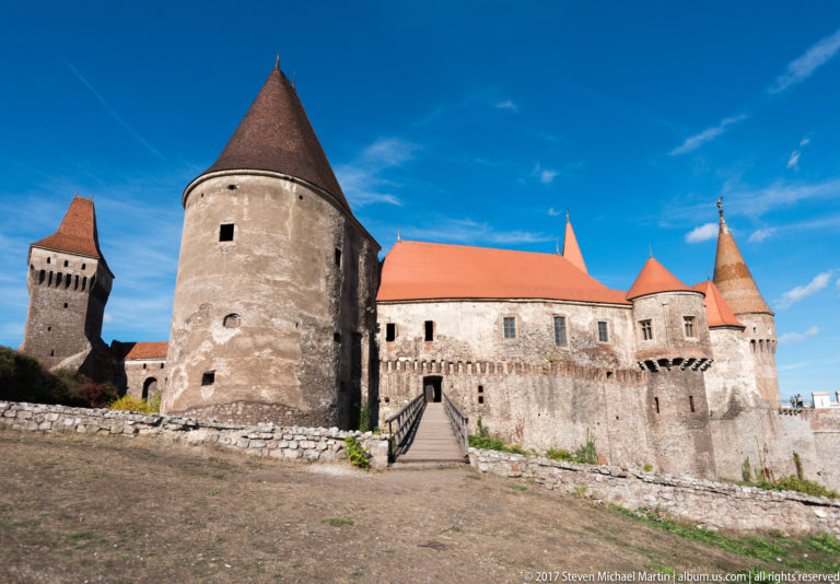 Broad exterior of Corvin Castle showing several towers, red roofs, long stone walls, and the entrance bridge.