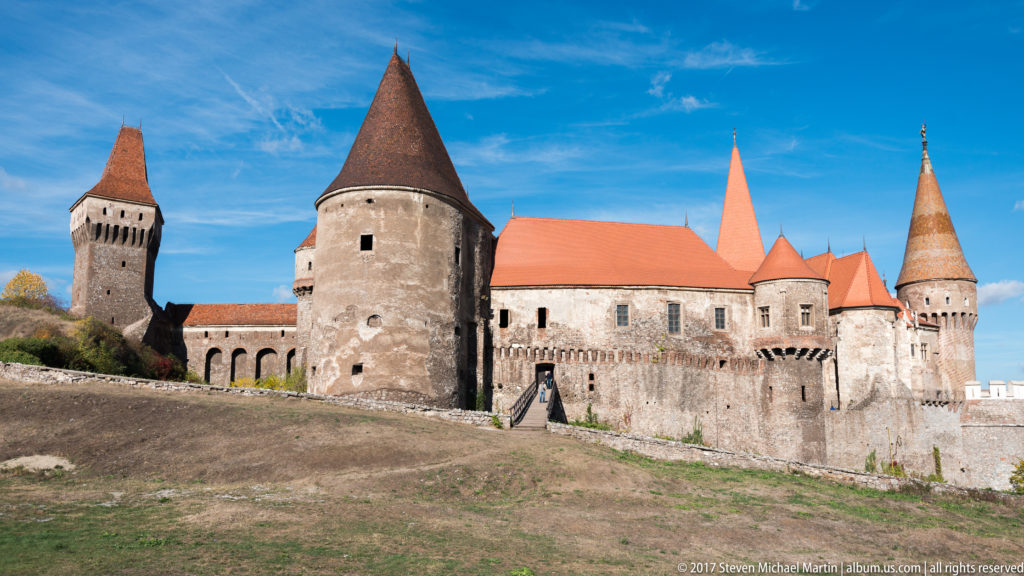 Wide side view of Corvin Castle with square and round towers, red roofs, and long stone wings.