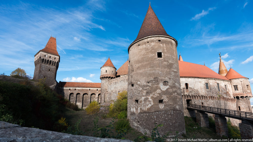Round tower, square tower, arcaded wall, and access bridge at Corvin Castle above the ravine.