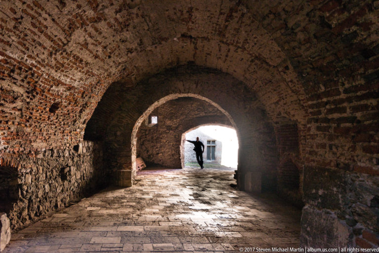 Brick-vaulted passage with worn stone floor and a visitor silhouetted at the bright far opening.