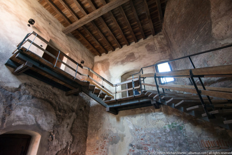 Modern metal-and-timber stairs crossing rough historic masonry beneath exposed roof beams at Corvin Castle.