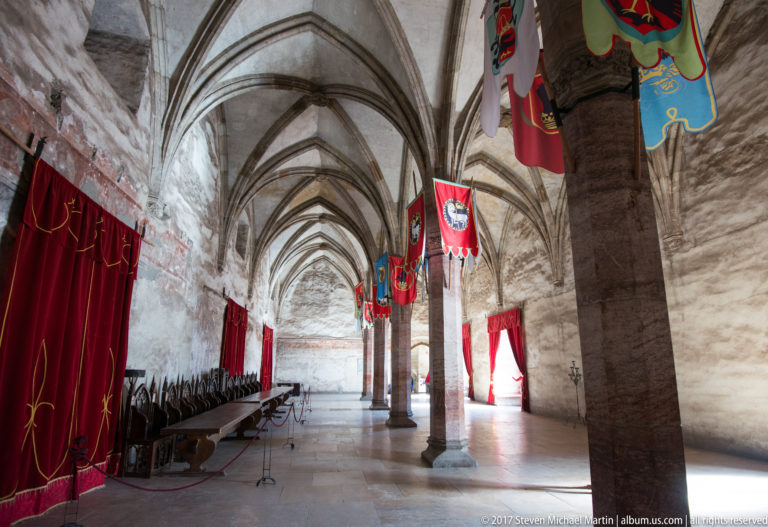 Long Gothic hall with rib vaults, stone columns, heraldic banners, red curtains, and seating along one wall.
