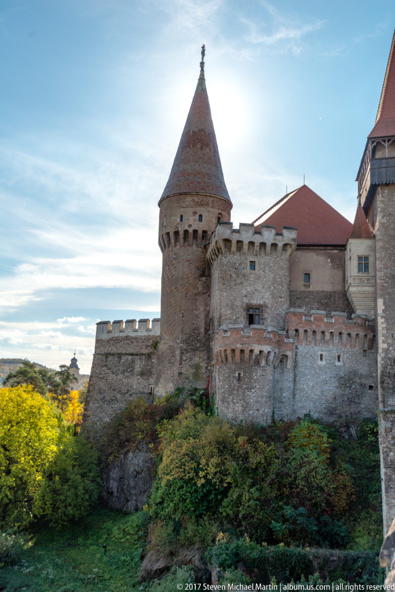 Round stone tower with conical roof and projecting defensive masonry above vegetation at Corvin Castle.