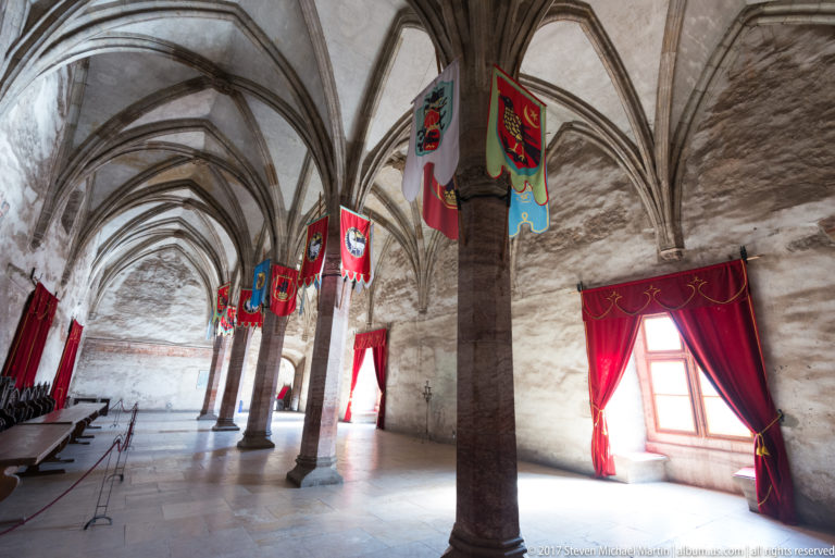 Rib-vaulted hall with heraldic banners and a red-curtained window at Corvin Castle.
