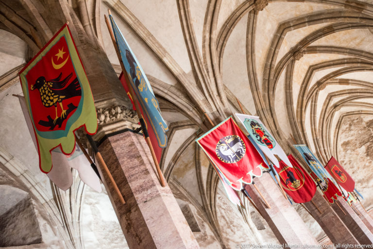Colorful heraldic banners hanging between stone columns and rib vaults in Corvin Castle.