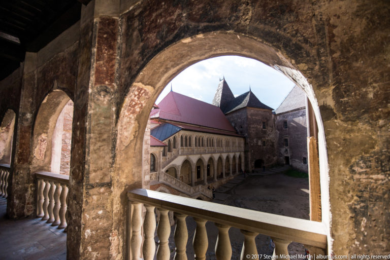 Inner courtyard framed by a large stone arch and balustrade, with arcaded galleries and red roofs beyond.
