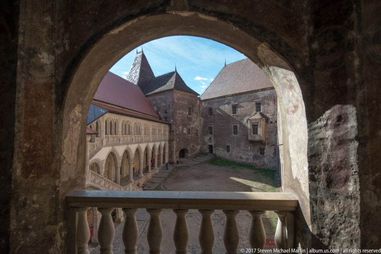 Courtyard seen through weathered stone arch and balustrade, with arcades and steep roofs beyond.