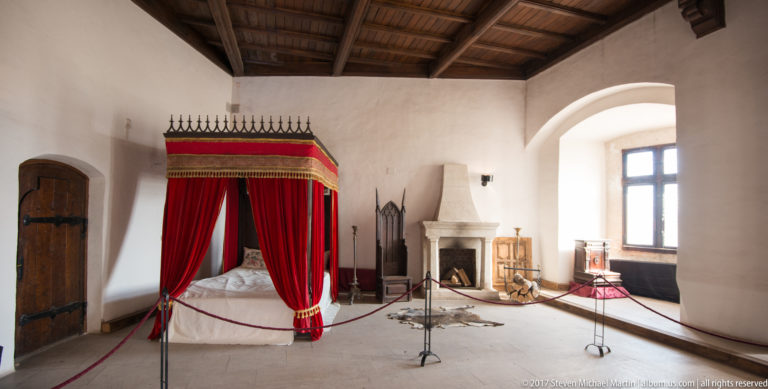 Red-canopied bed beside a fireplace in a white-walled castle room with timber ceiling.