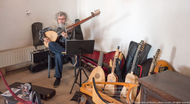 White-haired bearded musician playing a plucked string instrument beside several displayed lutes and cases.