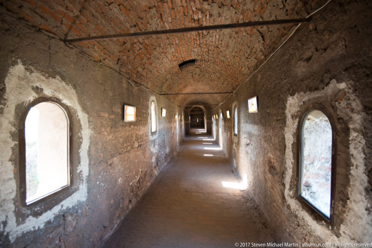Long narrow castle corridor with brick-vaulted ceiling, rough walls, and repeated windows on both sides.