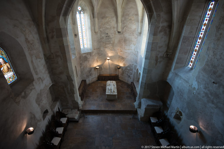 Small stone chapel with altar, tall stained-glass windows, pointed openings, and simple furnishings.
