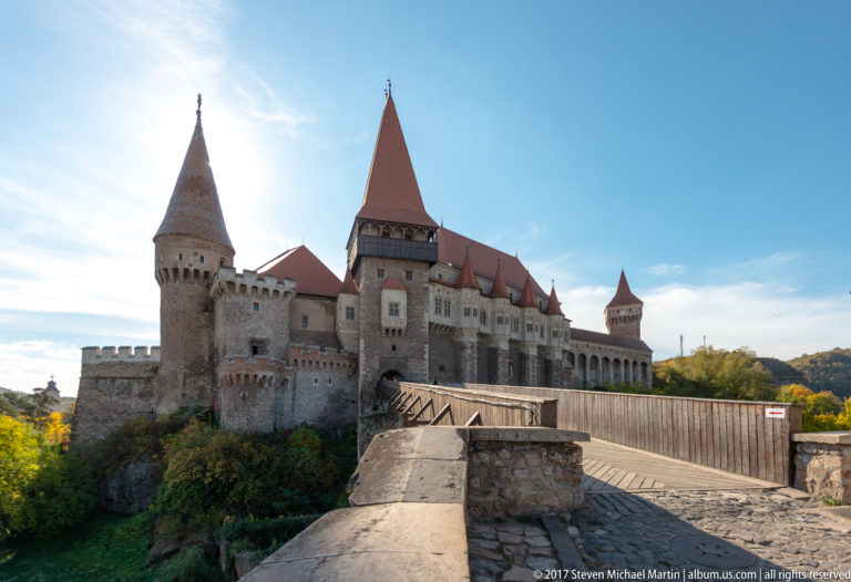 Wide approach to Corvin Castle with bridge, central gate tower, round tower, and long red-roofed wing.