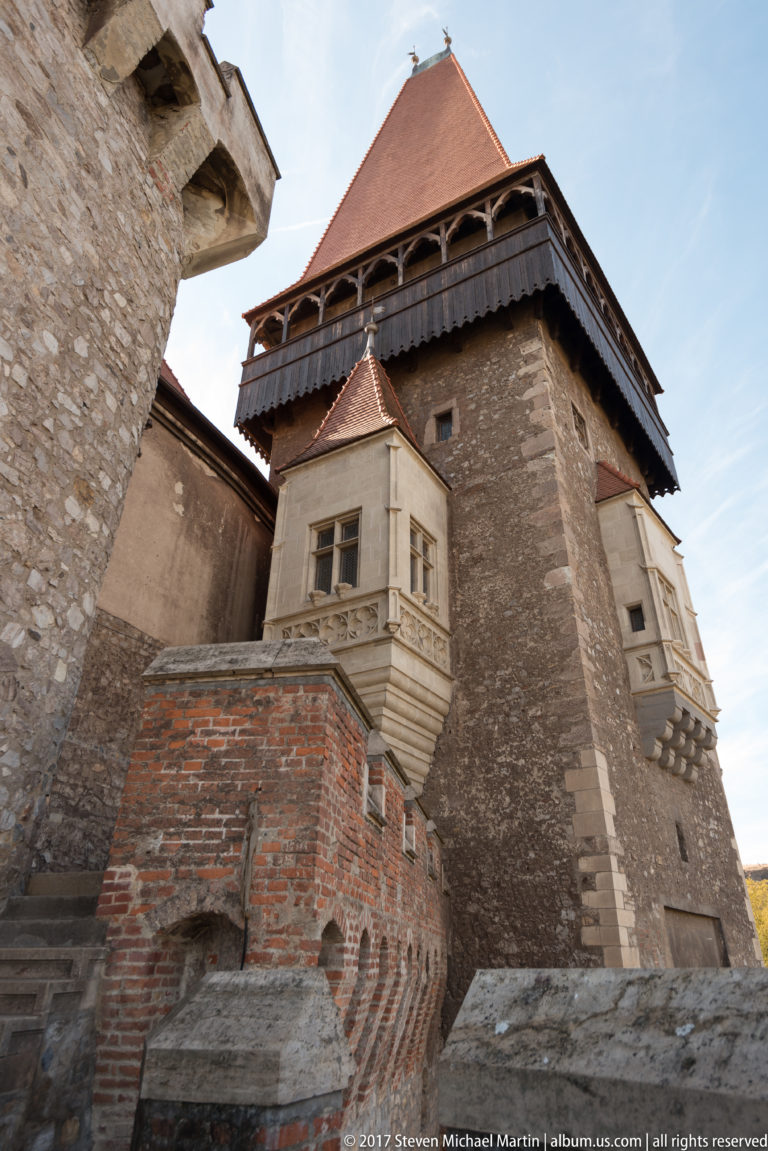 Tall castle wing with timber upper gallery, red roof, projecting stone window bay, and lower brickwork.
