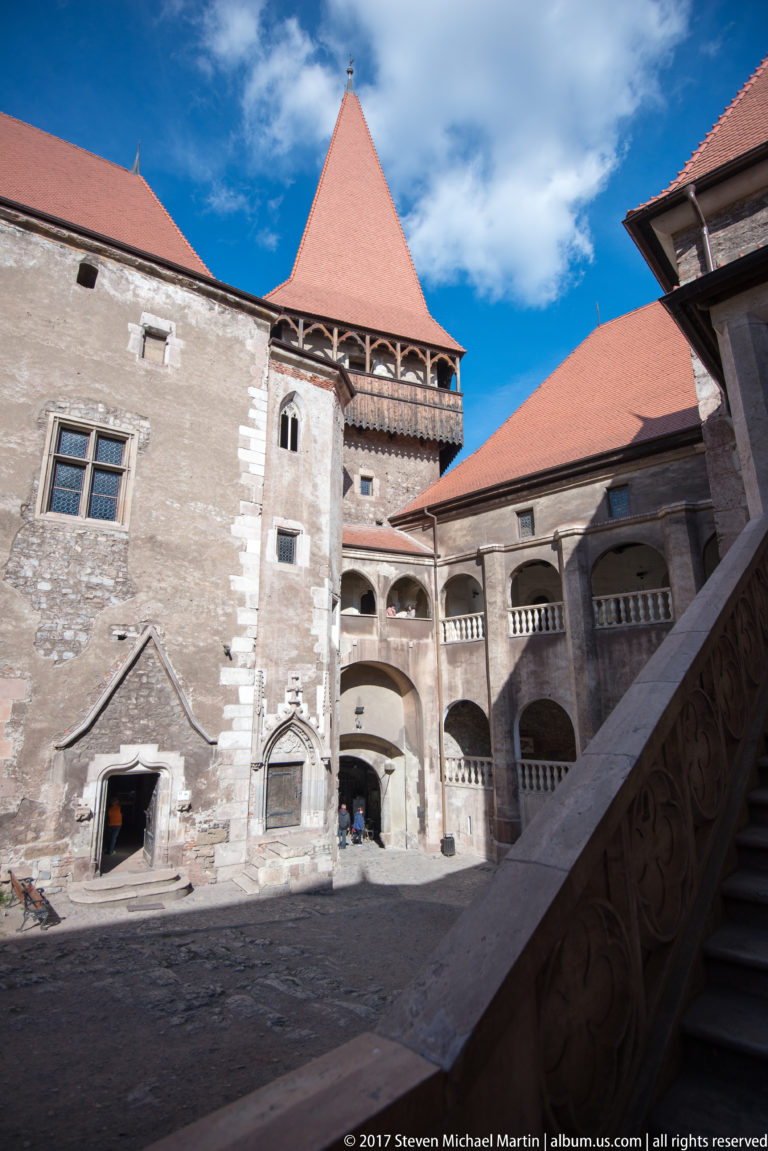 Corvin Castle courtyard with arcaded galleries, red-tiled roofs, weathered stone walls, and a broad exterior staircase.