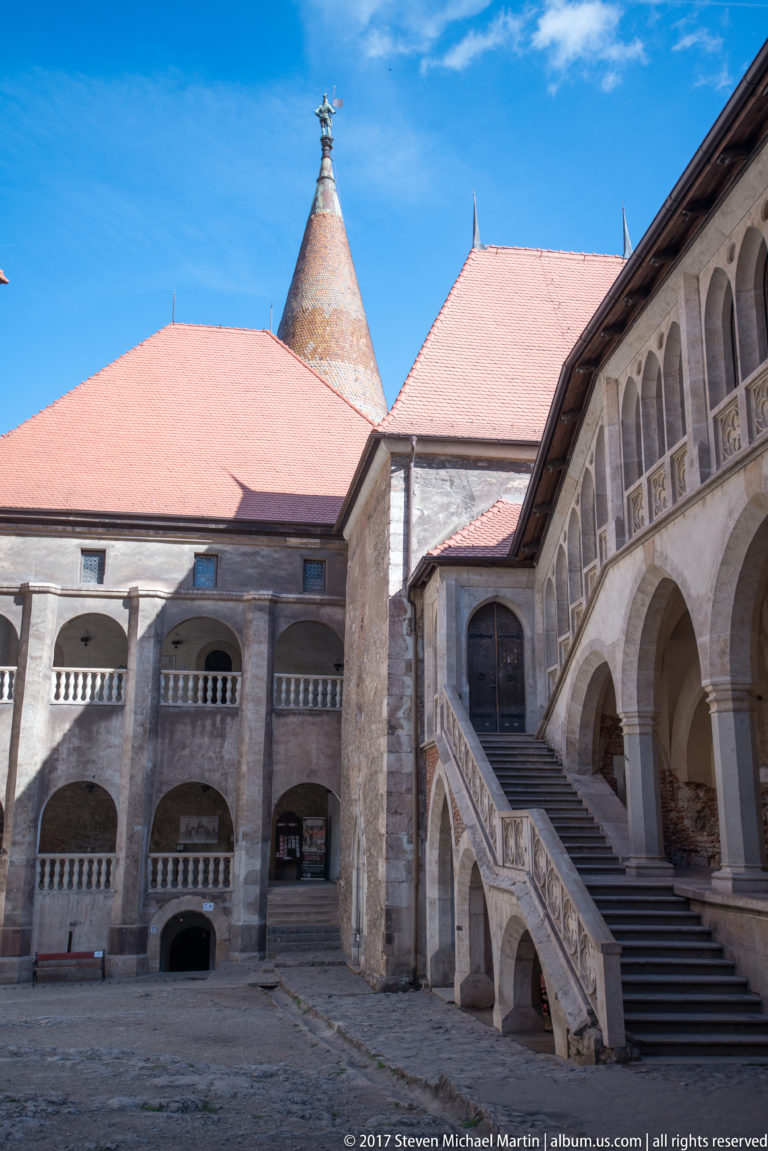 Arcaded Corvin Castle courtyard with monumental stone staircase, carved balustrade, red roofs, and conical tower roof.