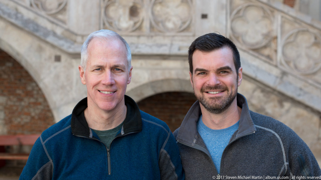 Two smiling male visitors posing together before carved stonework and an arched opening in Corvin Castle’s courtyard.