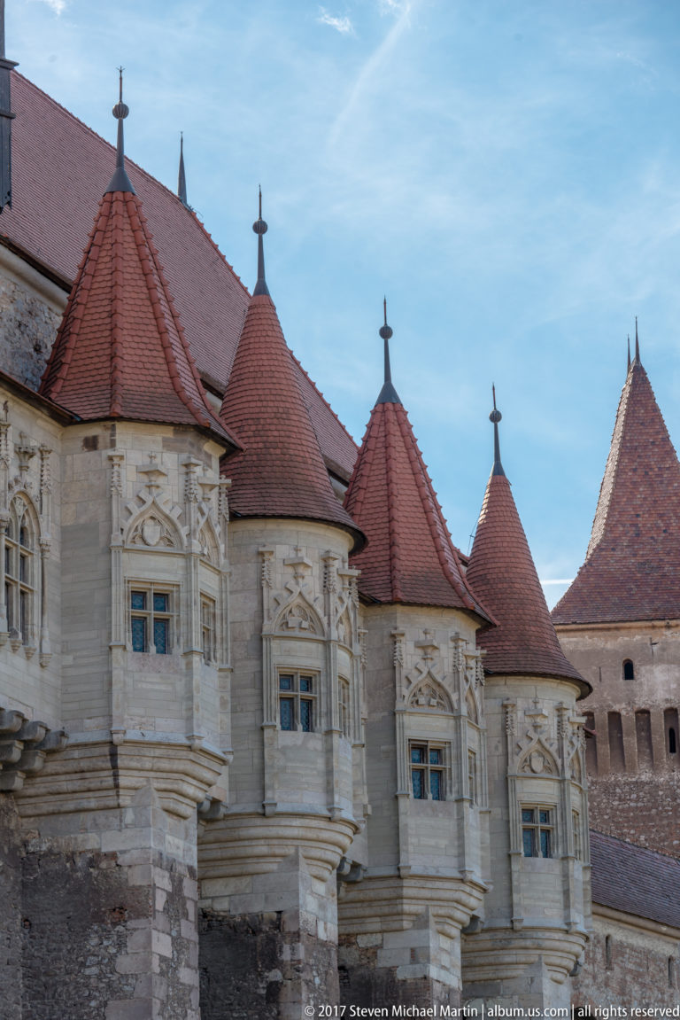 Ornate projecting stone window bays with carved decoration beneath steep red-tiled roofs on Corvin Castle’s residential façade.