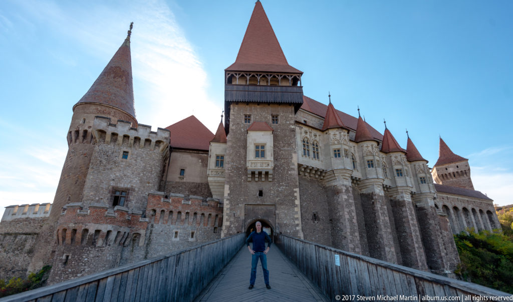 Visitor centered on Corvin Castle bridge beneath the tall gate tower and surrounding towers.
