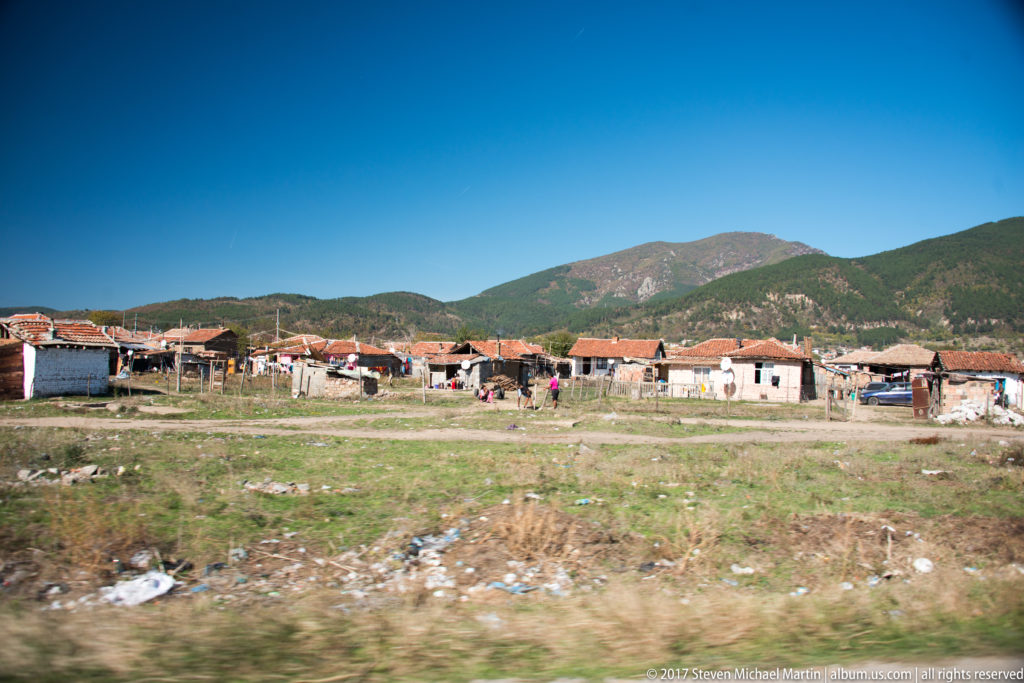 Wide residential settlement across rough open ground, backed by a broad forested mountain beneath a clear blue sky.