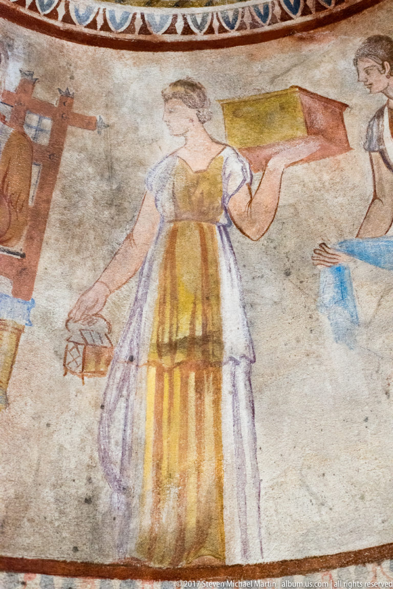 Painted woman in a striped ochre dress carrying a rectangular red-and-yellow box, with another figure partly visible at right.