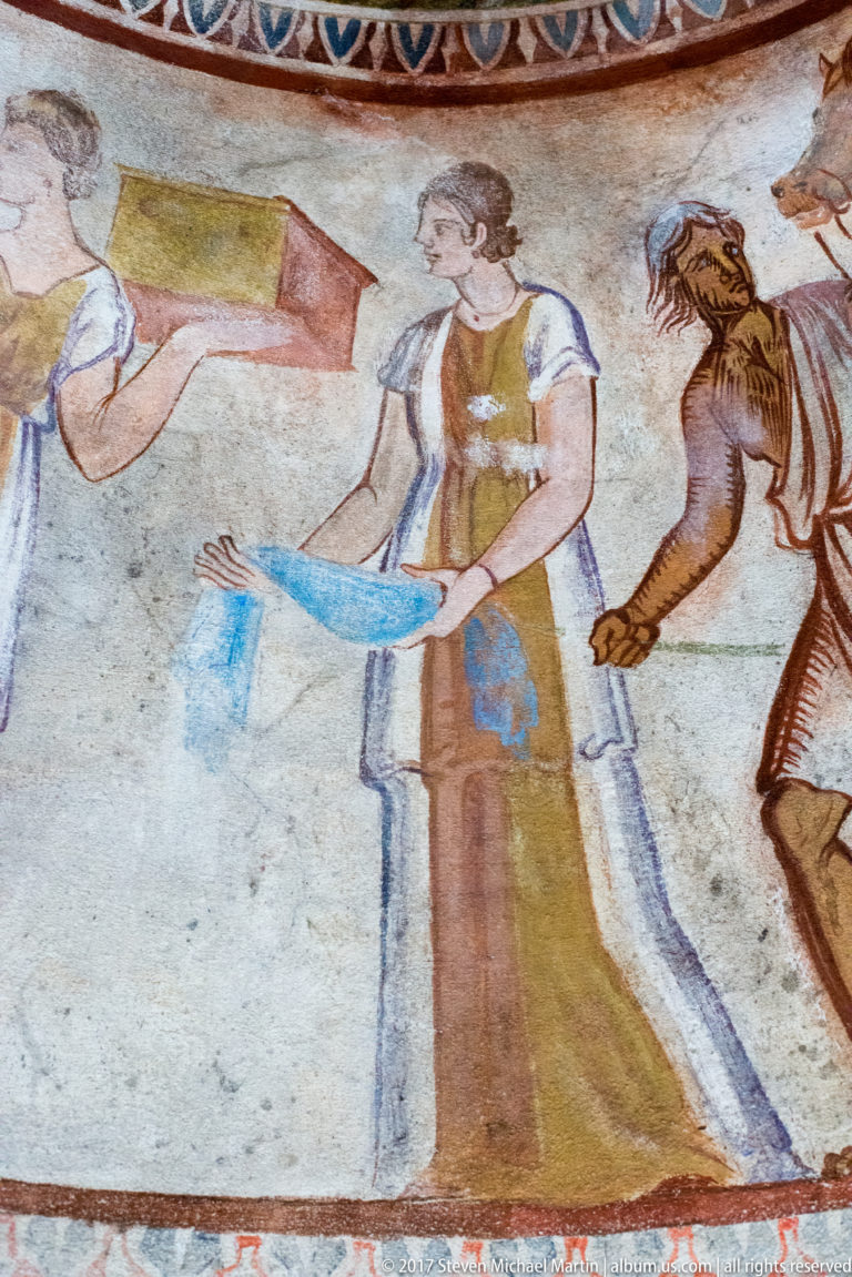 Painted woman in a long ochre-and-white garment carrying a bright blue object, with a darker male figure behind.