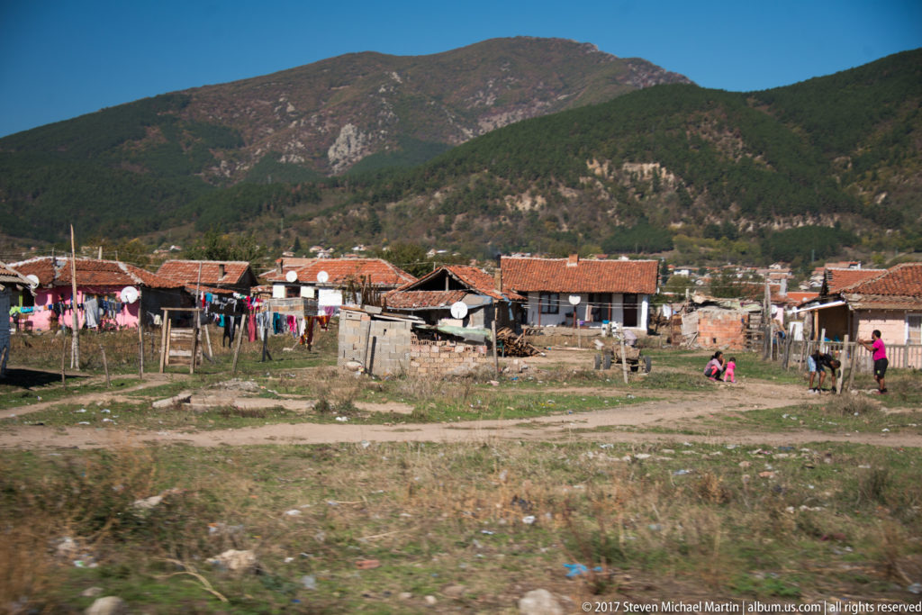 Low houses, fences, laundry, and residents in an open settlement beneath wooded mountains near Kazanlak.