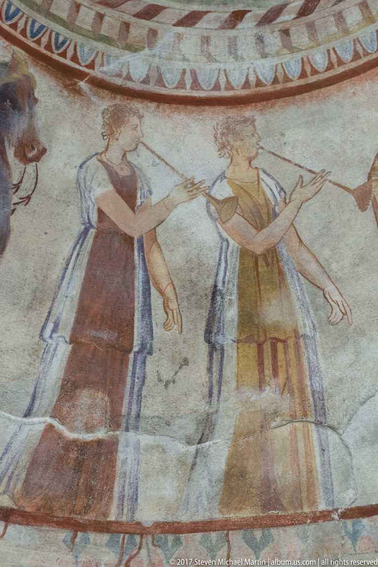 Two painted standing figures in long layered garments face each other and gesture beneath a patterned border.