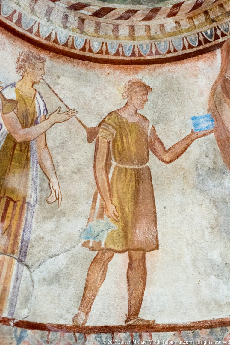 Two painted attendants in ochre and brown garments carrying bright blue vessels across a pale wall frieze.